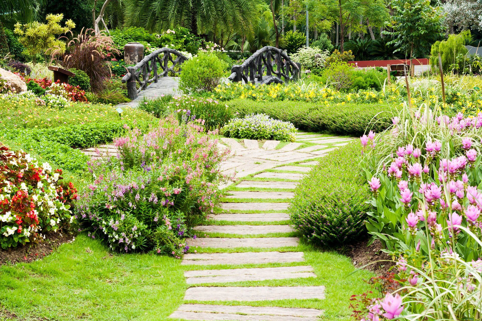 Gepflegter Garten mit Naturstein-Weg, bunten Blumenbeeten und rustikaler Holz-Brücke im Hintergrund